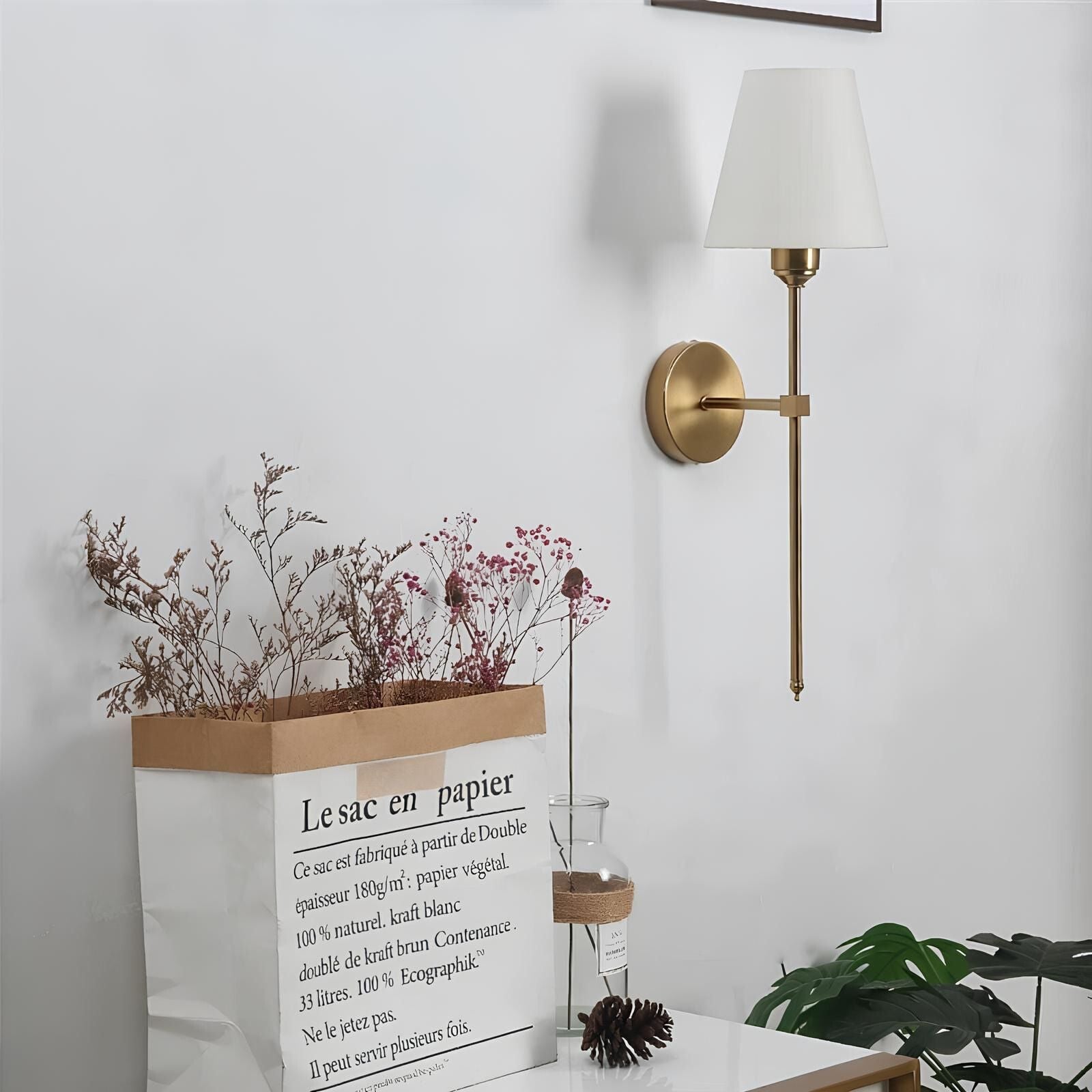 De Aurelia Wall Light in een klassieke gouden afwerking, verlicht een minimalistisch interieur met een elegante witte lampenkap. De lamp is geplaatst boven een decoratieve papieren zak met gedroogde bloemen en een stijlvolle vaas, wat zorgt voor een warme en verfijnde sfeer.