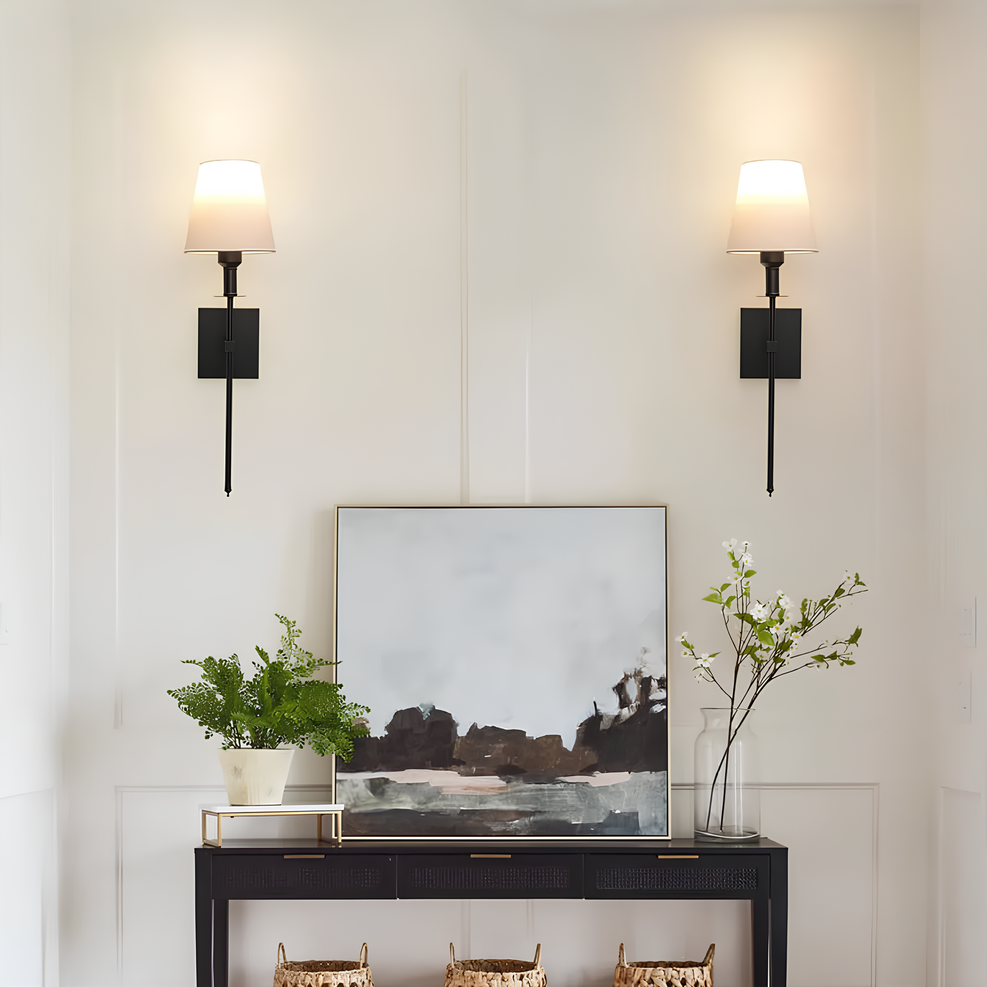 De Aurelia Wall Light in een elegante zwarte afwerking met een witte lampenkap verlicht een chique interieur. Twee wandlampen zijn gemonteerd naast een kunstwerk, met groene planten en decoratieve accessoires, waardoor een warme en stijlvolle sfeer ontstaat.