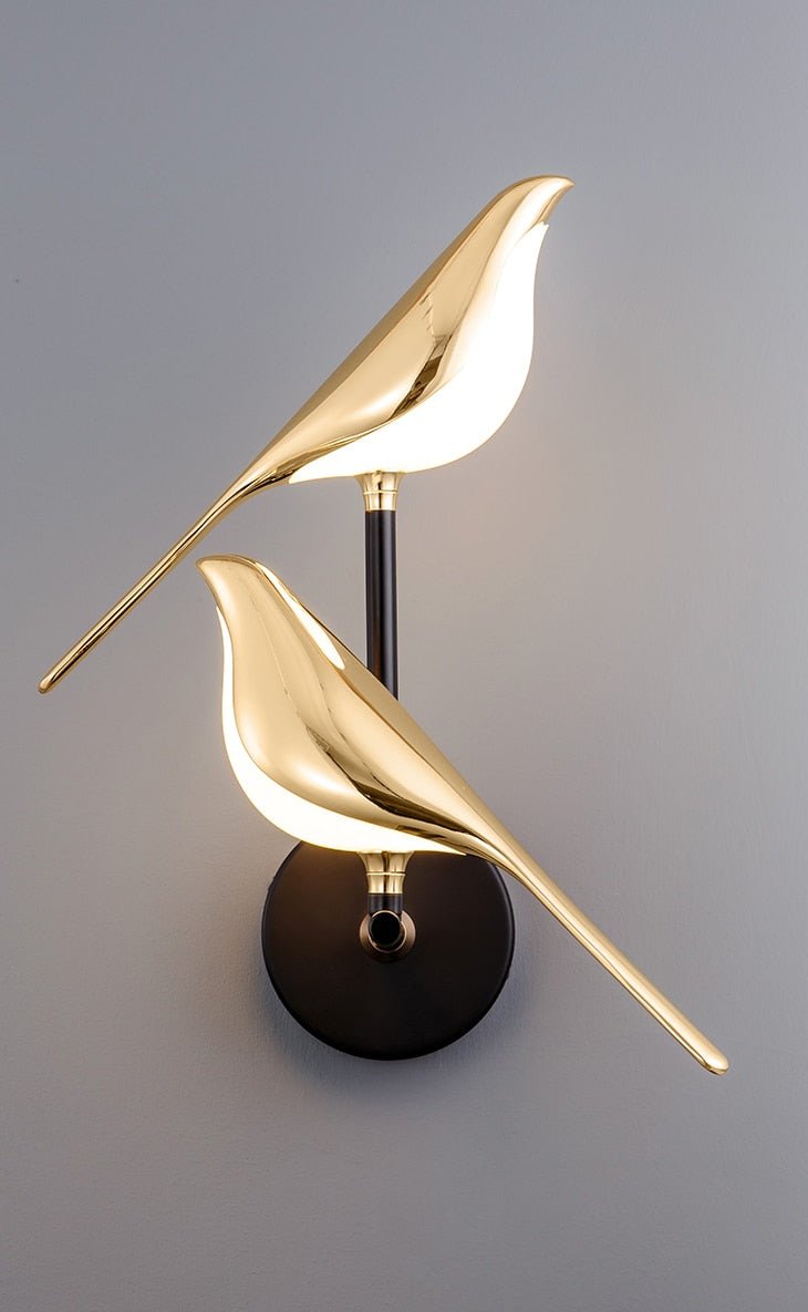 Auroria wandlamp met een dubbel vogelontwerp in glanzend goud, bevestigd op een zwarte wandbeugel, perfect voor een artistiek en sfeervol interieur.
