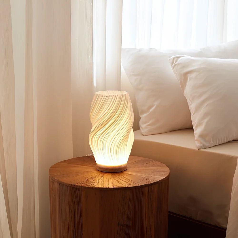 De Balaara Serene Wavecrest Lamp in een kalme slaapkamer, geplaatst op een houten tafel naast een bed met witte kussens, straalt zacht, warm licht uit dat een rustgevende sfeer creëert.