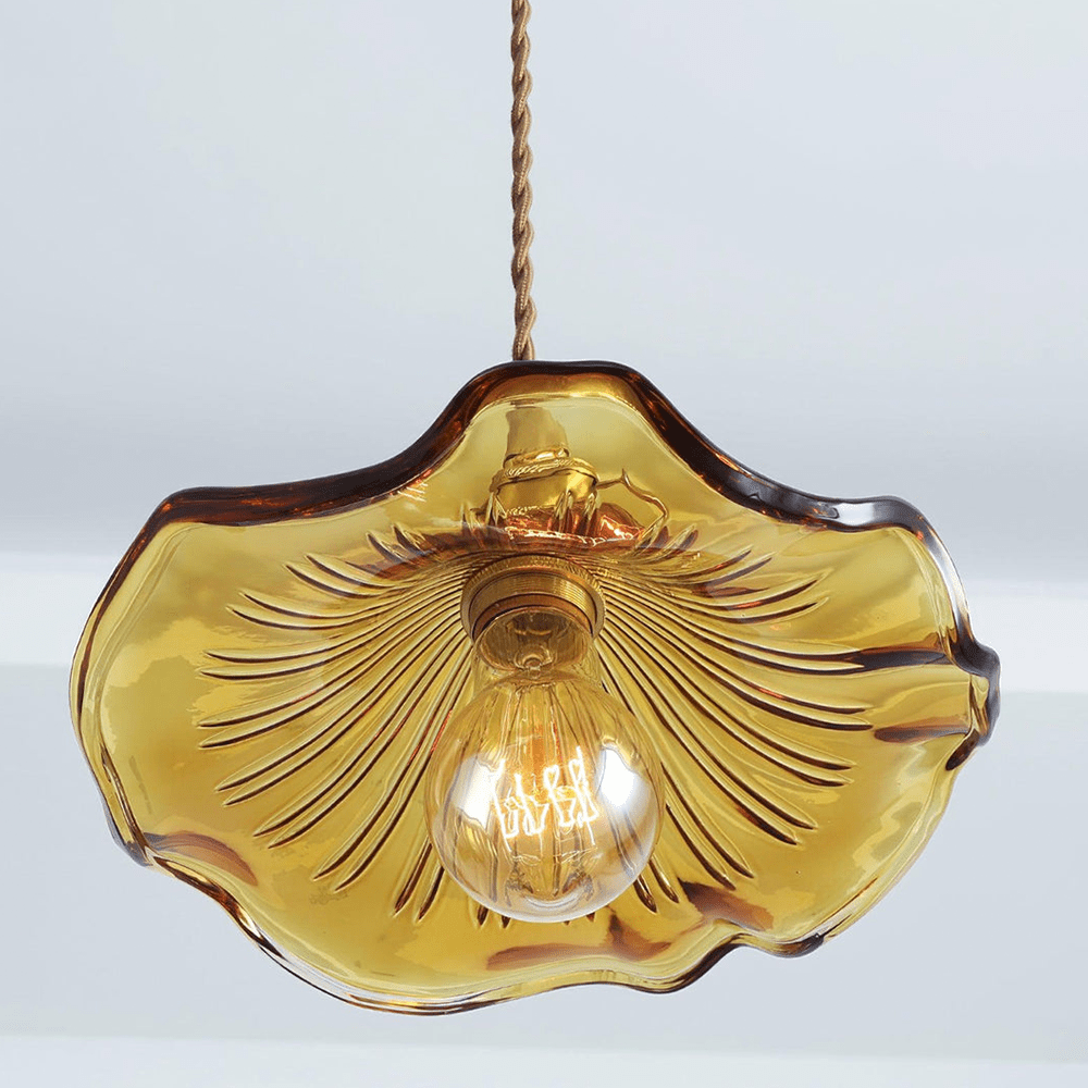 Close-up van de BloomLume hanglamp met een golvende amberkleurige glazen kap en een elegante gouden fitting. De warme gloed van het filamentlampje benadrukt het unieke ontwerp, ideaal voor het toevoegen van een stijlvolle en natuurlijke sfeer aan woonkamers, eetruimtes of creatieve interieurs.