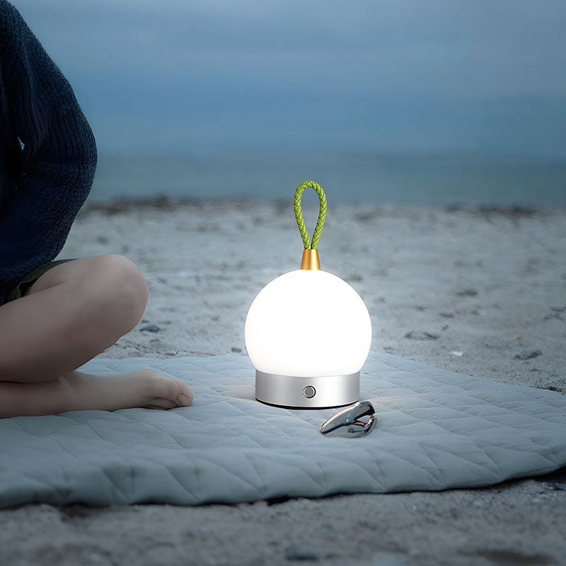 Caleo bedlamp met een bolvormig design en zilverkleurige basis, verlicht op een strandkleed bij zonsondergang. De lamp heeft een verstelbare groene lus aan de bovenkant, waardoor hij makkelijk te verplaatsen is. Ideaal voor buitengebruik of sfeervolle verlichting tijdens een avond aan het strand of in de tuin.