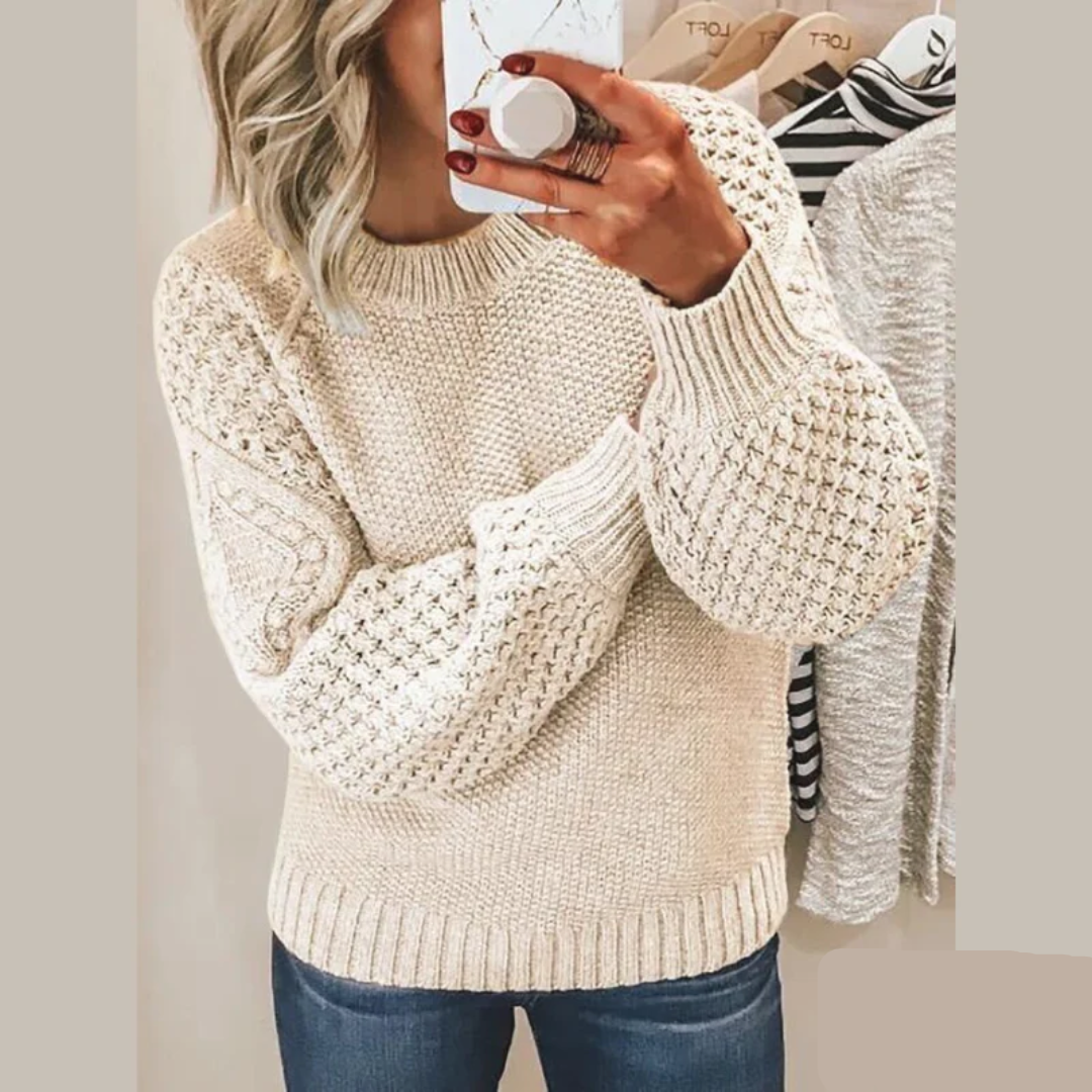 Een vrouw draagt een zachte, gebreide trui in een neutrale beige kleur met een mooi textuurpatroon op de mouwen en schouders. De trui heeft een klassieke ronde hals en biedt zowel comfort als stijl voor een casual look. Ze houdt een kop koffie vast terwijl ze zich in de spiegel bekijkt.