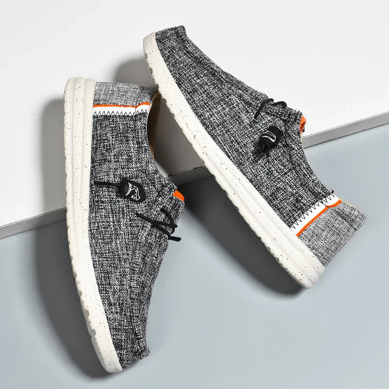 Lichte, grijze canvas slip-on schoenen voor heren met ademende stof en een flexibele, comfortabele zool. Deze moderne, casual schoenen zijn ontworpen voor dagelijks gebruik, met opvallende oranje details voor een sportieve uitstraling. Ideaal voor wandelingen en ontspannen activiteiten.