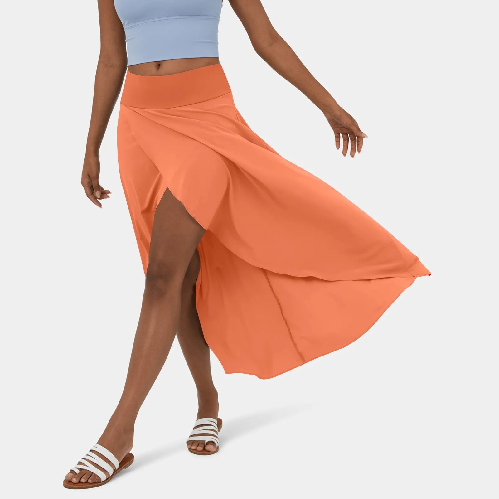 Een vrouw draagt een stijlvolle oranje rok die elegant meebeweegt met haar stappen. De rok heeft een vloeiende snit en een hoge taille, gecombineerd met een lichtblauw sporttopje en witte sandalen, perfect voor een casual en verfijnde zomerse look.