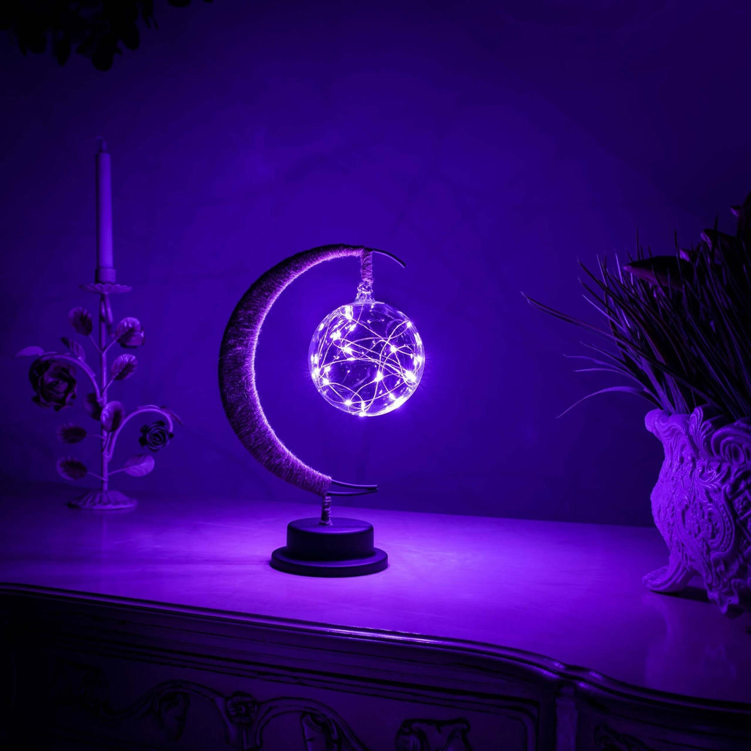 Een magische maanlamp verlicht met paarse LED-lichten, die een dromerige sfeer creëert. De lamp bevat een glinsterende bol van lichtjes, ondersteund door een elegante halve maanstructuur op een zwarte basis. De verlichting is perfect voor een mystieke, kalmerende ambiance in elke kamer.