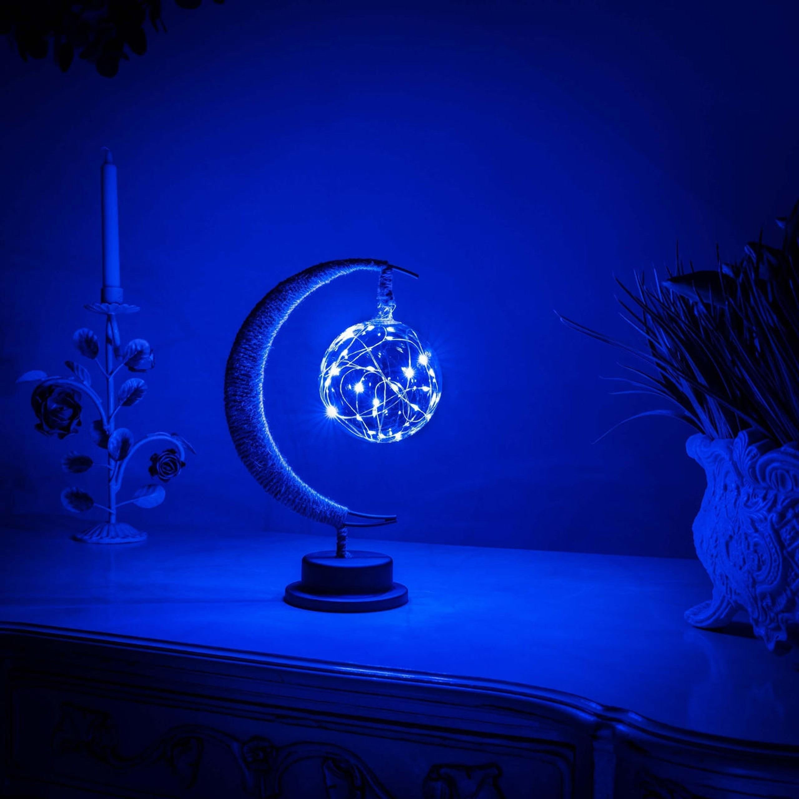 De GEMYNORA maanlamp, met een betoverende blauwe gloed, verlicht een nachtkastje in een rustige kamer. De lamp, met een halfmaansvorm en een stralende bol van lichtjes, creëert een magische sfeer en voegt een dromerige, kalmerende uitstraling toe aan het interieur.