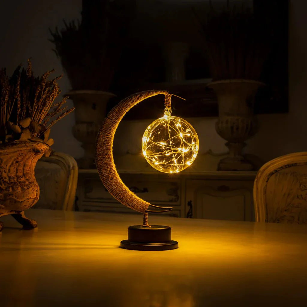 De GEMYNORA maanlamp straalt warm, magisch licht uit, met een elegante combinatie van een halfmaan en een bol van gloeiende draadjes. De lamp staat op een houten tafel, verlicht door het sfeervolle licht dat een rustgevende ambiance creëert in de kamer. Perfect voor het toevoegen van een betoverende touch aan elk interieur.