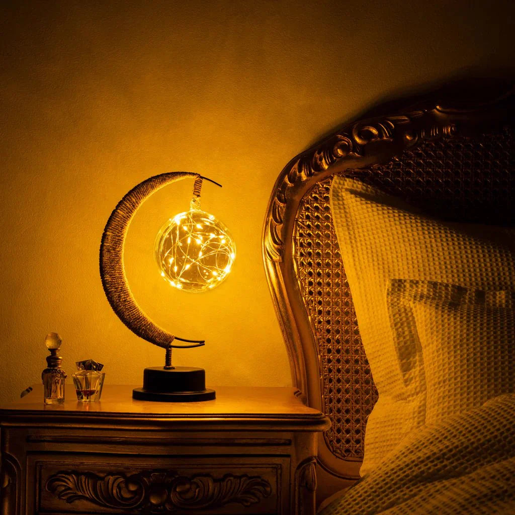 De GEMYNORA maanlamp, die een warme en betoverende gloed uitstraalt, staat op een nachtkastje naast een bed. De lamp, met een elegante maanvorm en een bol van lichtsnoeren, creëert een magische sfeer in de kamer, perfect voor ontspanning en romantische avonden.