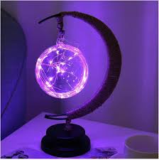 De GEMYNORA maanlamp verlicht met een betoverende paarse gloed, met een verlichte bol van lichtsnoeren die zachtjes schijnt. De lamp is geplaatst op een elegant zwart basis, met een maanvormige steun die een magische sfeer toevoegt aan elke kamer.