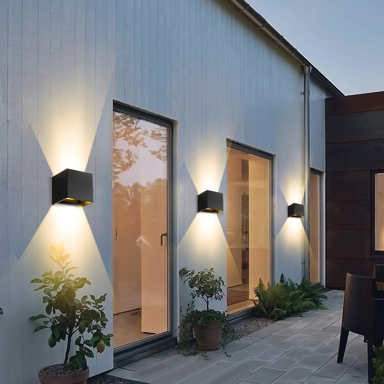 Een set moderne LED-wandlampen met een strak zwart vierkant ontwerp, gemonteerd op de buitenmuur van een huis, die stijlvol licht in twee richtingen uitstralen. Het warme licht benadrukt de planten en ramen en creëert een uitnodigende en eigentijdse sfeer.