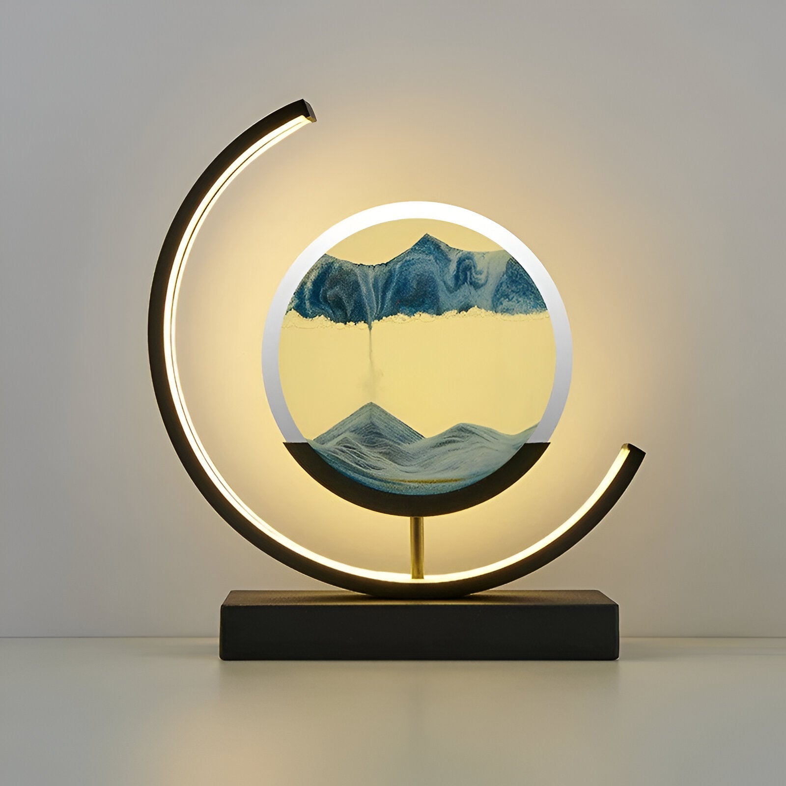 De GlareGlow Sandlamp is een kunstzinnige lamp met een veranderend zandpatroon in blauw en geel, omgeven door een elegante zwart-gouden basis. De lamp creëert een sfeervolle, rustgevende atmosfeer door de zachte verlichting die van de zijkanten uitstraalt. Het moderne design maakt deze lamp ideaal voor het verfraaien van elk interieur.
