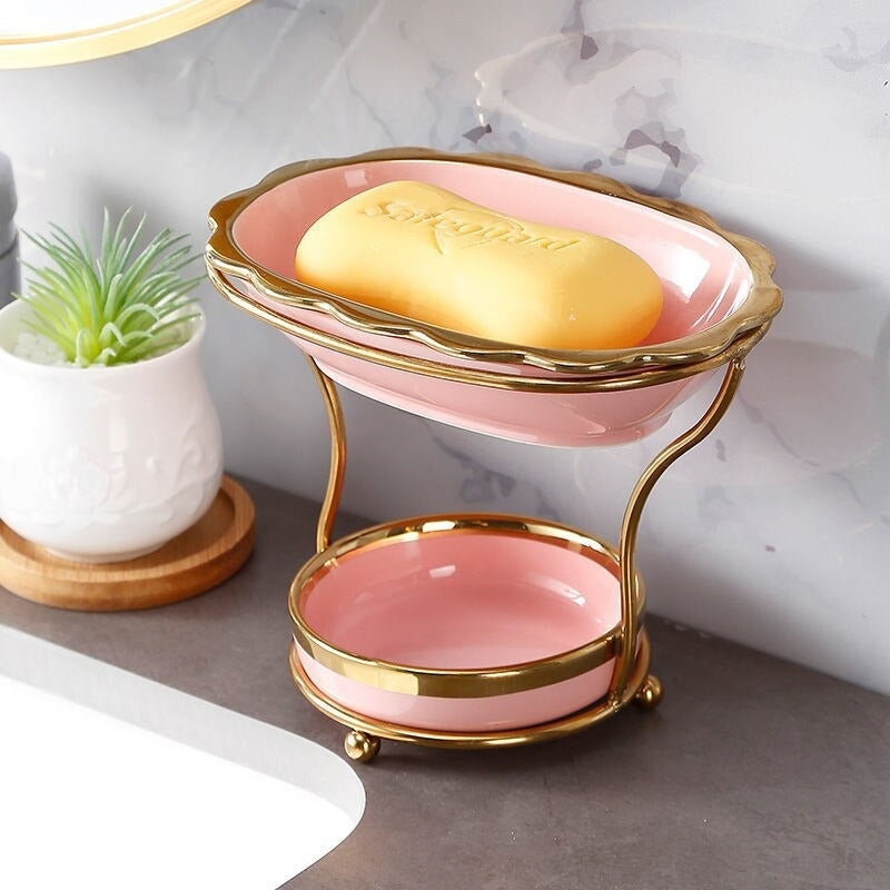 GleamEdge zeepbakje in zachtroze met gouden accenten, geplaatst op een elegante tweelaagse metalen standaard. Dit moderne en praktische ontwerp houdt zeep droog en voegt een luxe uitstraling toe aan elke badkamer.