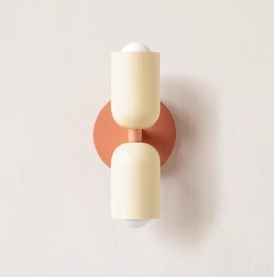 Een minimalistische wandlamp in Deense stijl met twee zachte, crèmekleurige lampenkappen en een warme oranje wandbeugel. De lamp heeft een strak en modern ontwerp, perfect voor het toevoegen van een subtiele, elegante verlichting aan elke kamer.