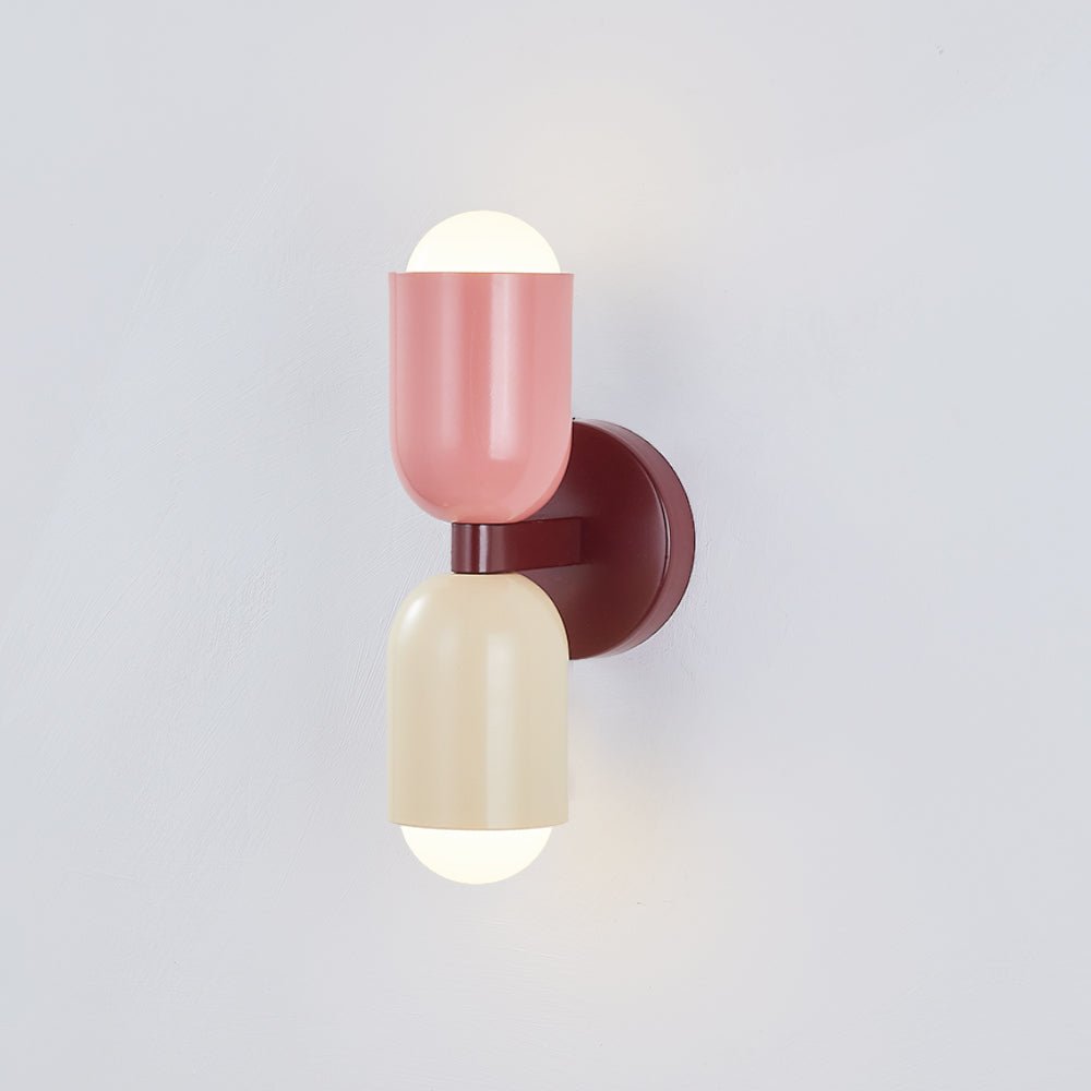 Een stijlvolle Glemlux wandlamp in Deense stijl, met een roze en crèmekleurige cilindervormige kap en een warme lichtbron. De lamp is gemonteerd op een slanke, bruine ronde basis en voegt een moderne en speelse uitstraling toe aan elke eigentijdse ruimte.