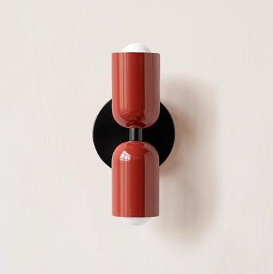 Een moderne Deense wandlamp met een elegant design, bestaande uit twee cilindervormige lampenkappen in een warme terracotta kleur, gemonteerd op een zwart muurbeugel. De lampen stralen een zachte, warme gloed uit en voegen een stijlvol en minimalistisch accent toe aan de ruimte.