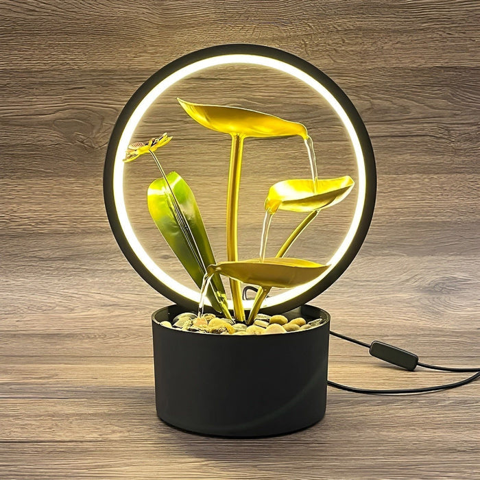 De Harmonia tafellamp, een stijlvolle en veelzijdige verlichting met een cirkelvormige LED-lamp, gouden bladeren en een rustgevende waterfunctie. De zwarte basis en natuurlijke details zoals kiezelstenen maken het een perfect decoratief stuk voor een modern interieur.
