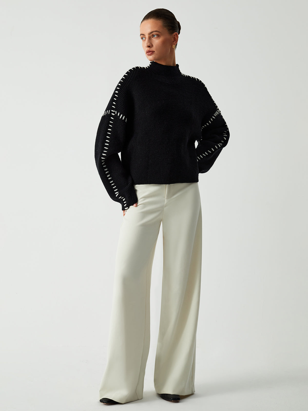 Model poseert in een Helvano oversized trui in zwart met witte stikseldetails langs de schouders en mouwen. De trui heeft een hoge kraag en een losse, comfortabele pasvorm. Gecombineerd met wijde, lichte broeken voor een moderne en elegante uitstraling. Perfect voor een casual maar stijlvolle look.