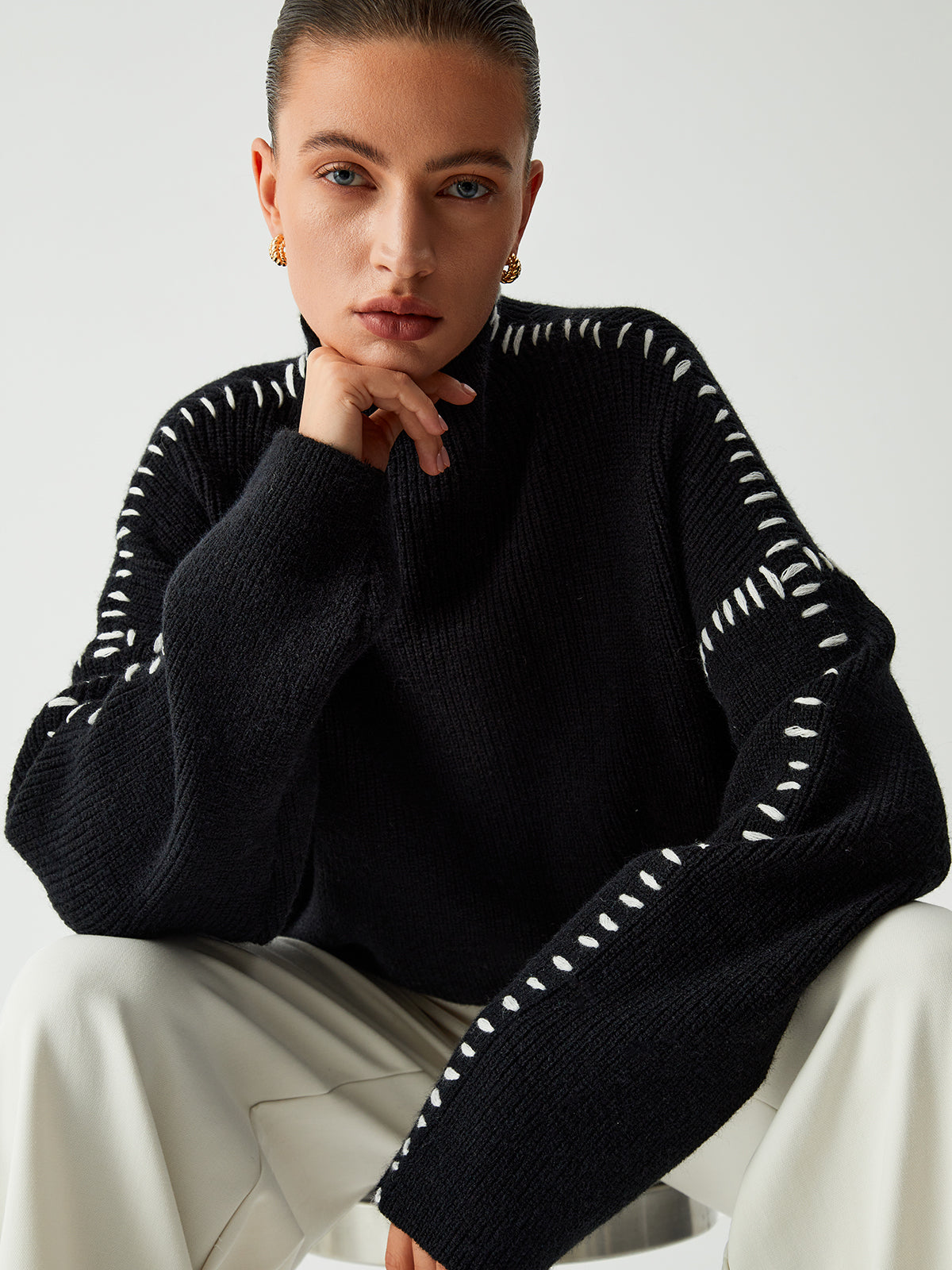 Model draagt een Helvano oversized trui in zwart met witte stikseldetails langs de mouwen en schouders. De trui heeft een hoge kraag en een relaxte pasvorm. Perfect voor een moderne en comfortabele winterlook. Gecombineerd met lichte broeken voor een stijlvol contrast.