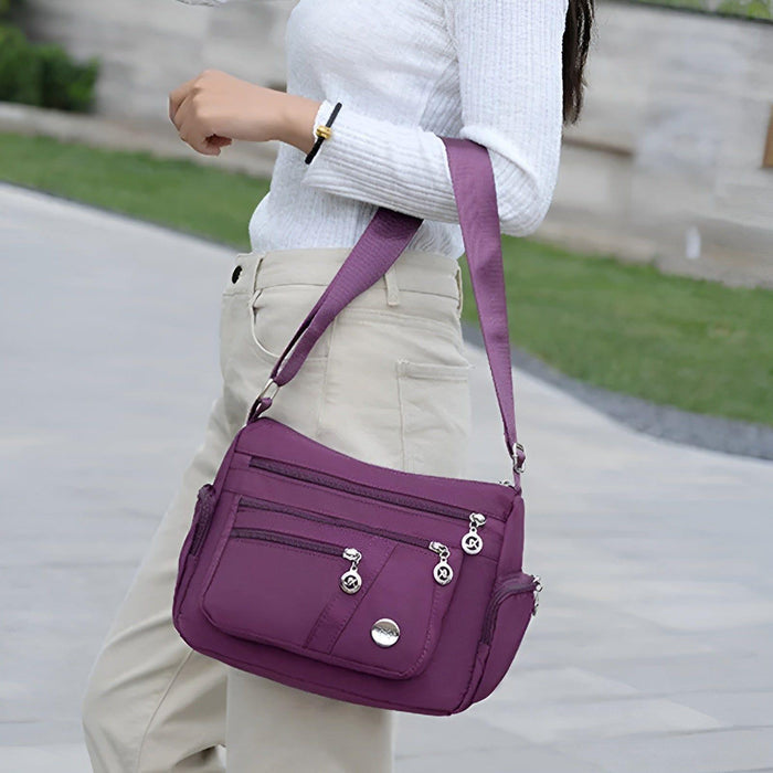 Vrouw met lange donkere haren draagt een stijlvolle paarse schoudertas met meerdere vakken en zilverkleurige ritssluitingen. De tas heeft een praktisch en veelzijdig ontwerp, perfect voor dagelijks gebruik of een dagje uit. De verstelbare schouderriem zorgt voor extra draagcomfort en flexibiliteit.