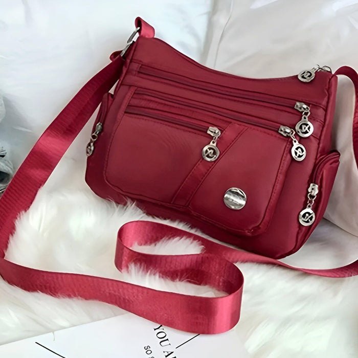 Stijlvolle rode schoudertas met meerdere vakken en zilverkleurige ritssluitingen, liggend op een wit oppervlak. De tas heeft een praktisch en veelzijdig ontwerp, ideaal voor dagelijks gebruik. De verstelbare schouderriem zorgt voor extra draagcomfort en flexibiliteit.