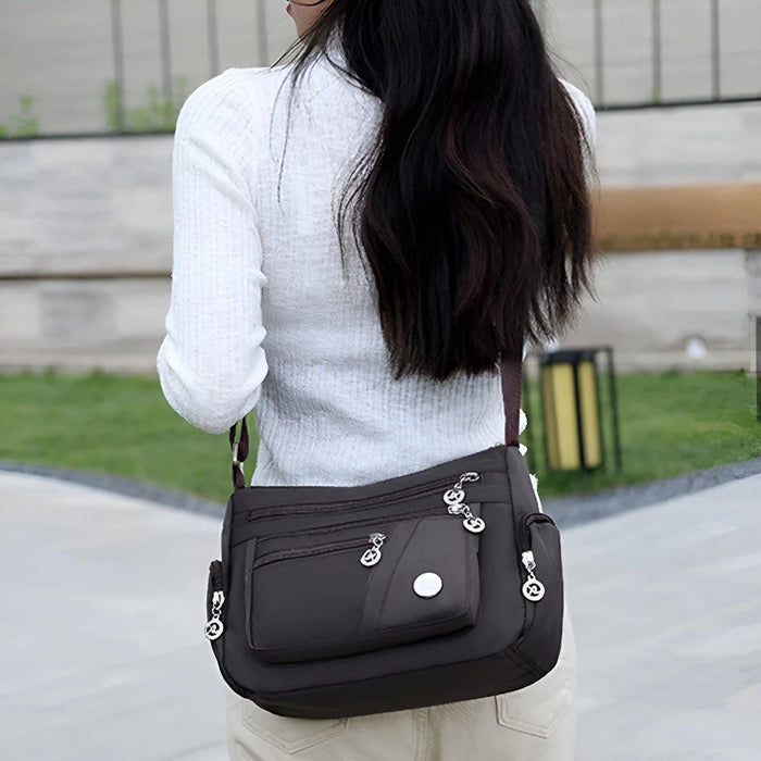 Vrouw met lange donkere haren draagt een stijlvolle zwarte schoudertas met meerdere vakken en zilverkleurige ritssluitingen. De tas heeft een praktisch en veelzijdig ontwerp, ideaal voor dagelijks gebruik of een dagje uit. De verstelbare schouderriem zorgt voor extra draagcomfort.