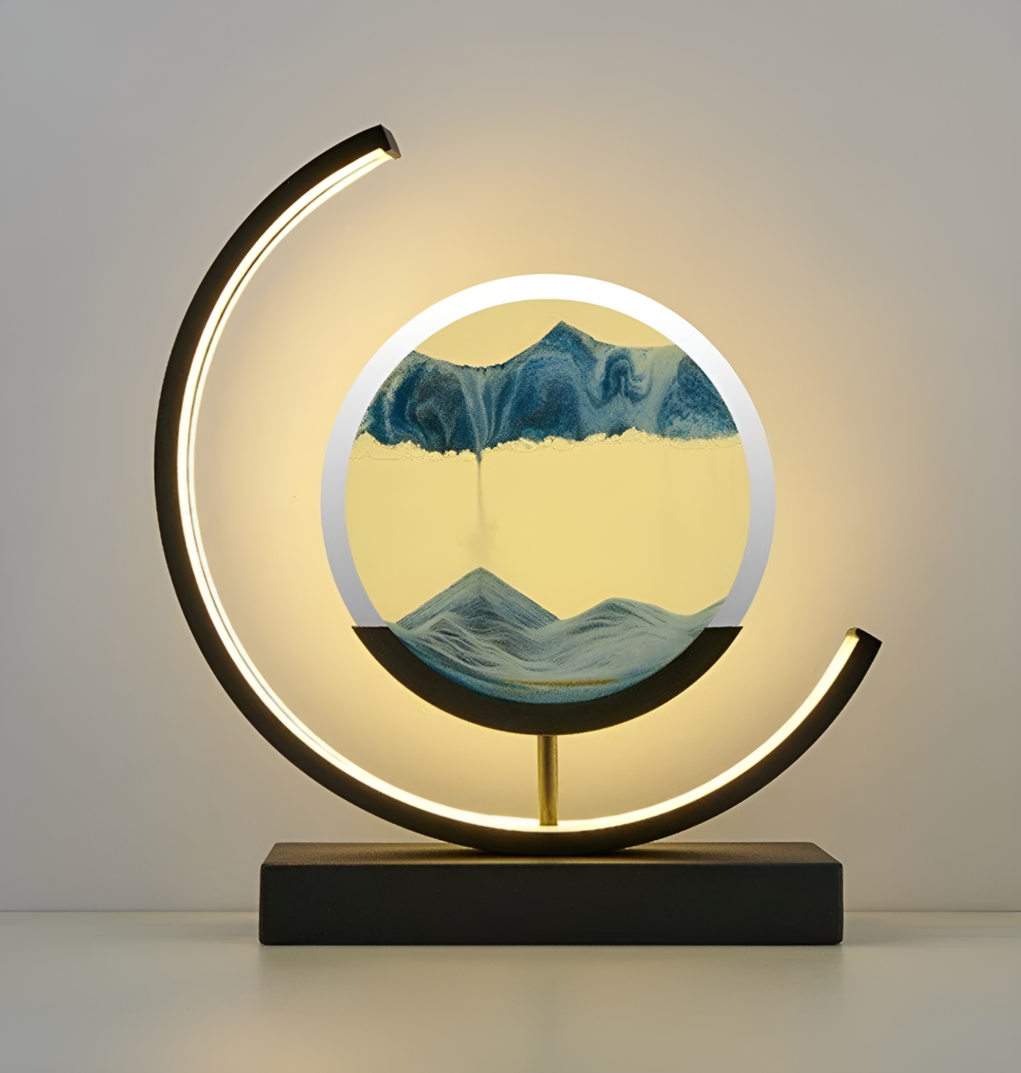 De afbeelding toont een moderne LED-lamp met een ronde basis en zandkunst in het midden, wat een kalmerend, dynamisch effect creëert. De lamp is verlicht en werpt zacht licht op de paarse en gele tinten in het zand. De lamp heeft een strak, minimalistisch ontwerp, met een gebogen LED-licht rondom de zandkunst, waardoor het een elegante uitstraling aan elke ruimte geeft.