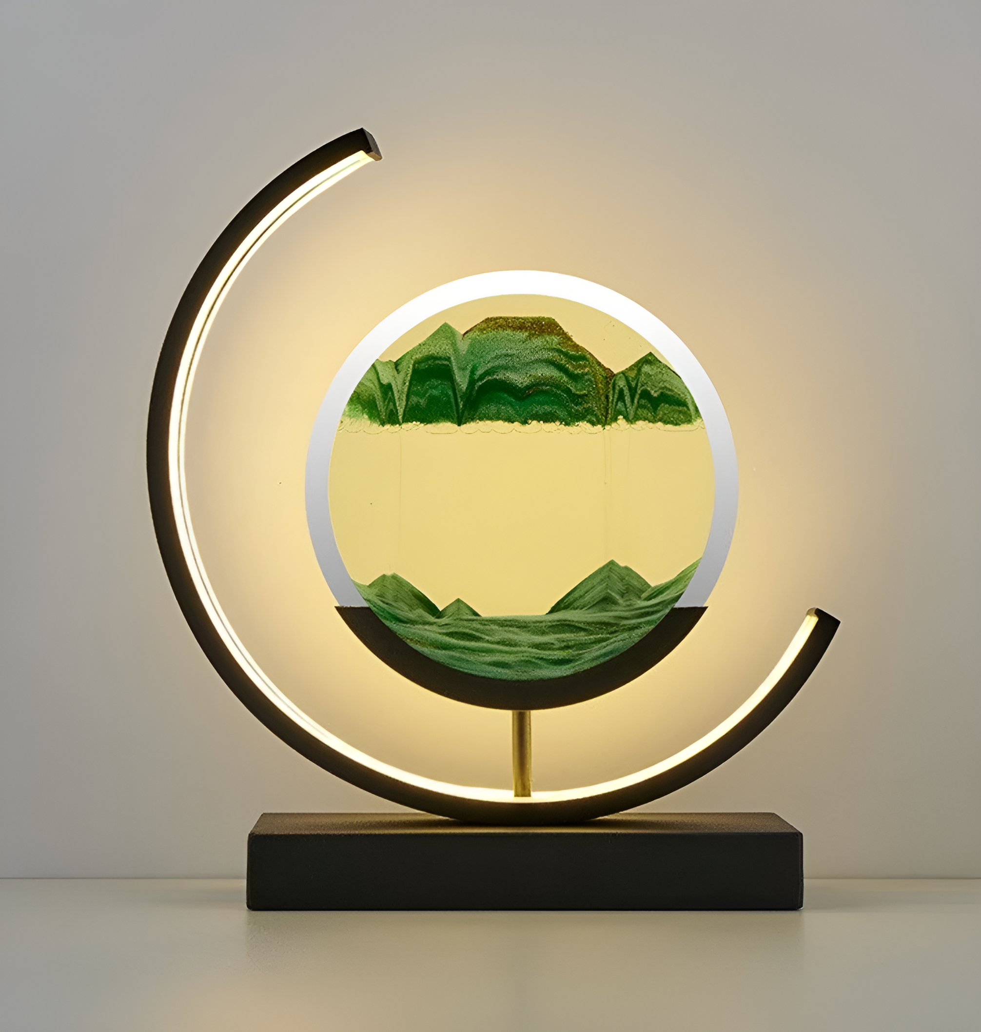 De afbeelding toont een elegante LED-lamp met een unieke ronde zandkunst in het midden. Het zand is in verschillende tinten groen en geel, wat een bergachtig landschap simuleert. De lamp wordt omlijst door een gebogen LED-licht dat een warme gloed uitstraalt, perfect voor het creëren van een sfeervolle en moderne ambiance. De zwarte basis van de lamp biedt een strakke en verfijnde afwerking, waardoor deze lamp een stijlvolle aanvulling is op elk interieur.