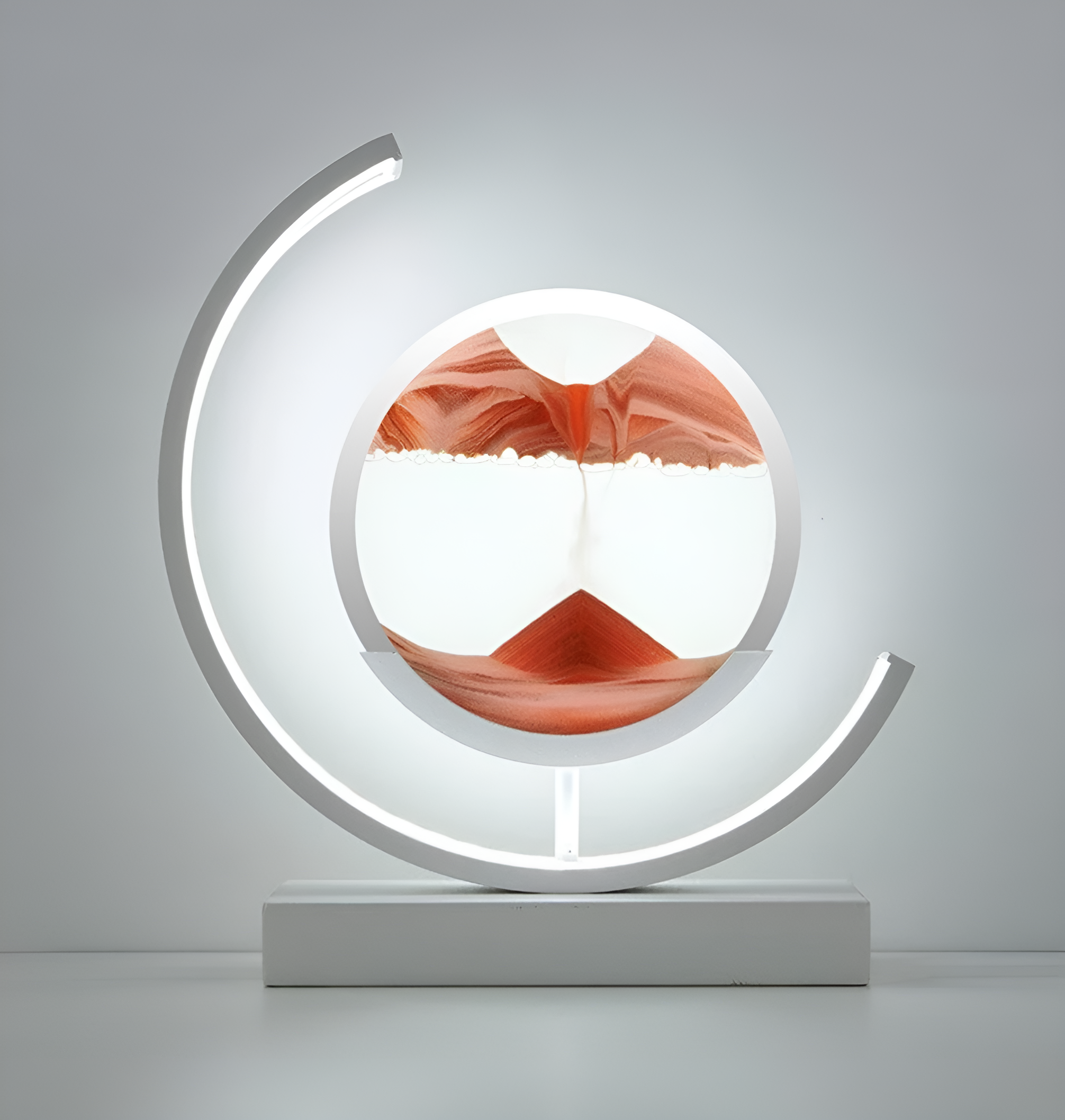 De afbeelding toont een stijlvolle LED-lamp met een uniek zandkunstontwerp in het midden. Het zand is verdeeld in zachte tinten van oranje en wit, die samen een bergachtig landschap creëren. De lamp wordt omlijst door een gebogen LED-licht dat een warme, uitnodigende gloed uitstraalt, perfect voor het creëren van een sfeervolle en moderne uitstraling. De strakke, witte basis maakt het geheel af, wat deze lamp tot een elegante toevoeging maakt voor elk interieur.