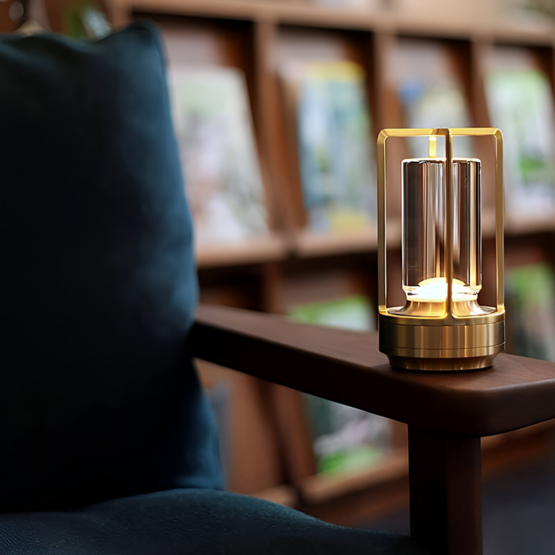 De afbeelding toont de Jovien Lamp, een elegante, waterbestendige lamp met een modern ontwerp. De lamp staat op de armleuning van een comfortabele stoel en straalt een warme gloed uit, wat een gezellige sfeer creëert. Achter de lamp is een boekenkast zichtbaar, wat de veelzijdigheid van de lamp in verschillende interieurinstellingen benadrukt. Het gouden frame van de lamp voegt een luxe uitstraling toe aan de ruimte.