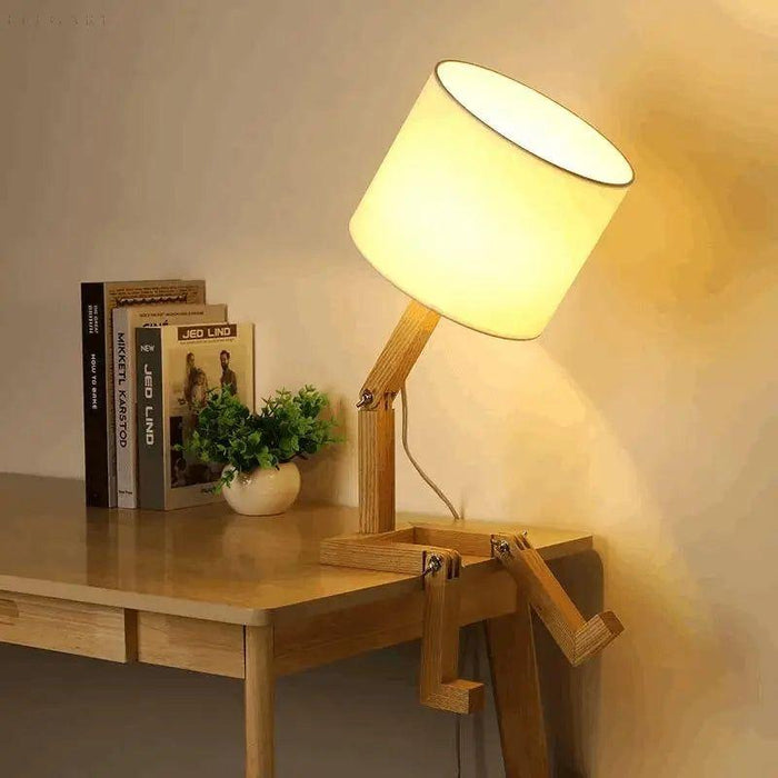 Klaarlicht tafellamp in de vorm van een zittend figuur op een houten bureau. De lamp heeft een houten frame met verstelbare 'armen' en 'benen', en een grote, witte lampenkap die warm licht verspreidt. Perfect voor een speelse en stijlvolle toevoeging aan elk interieur.