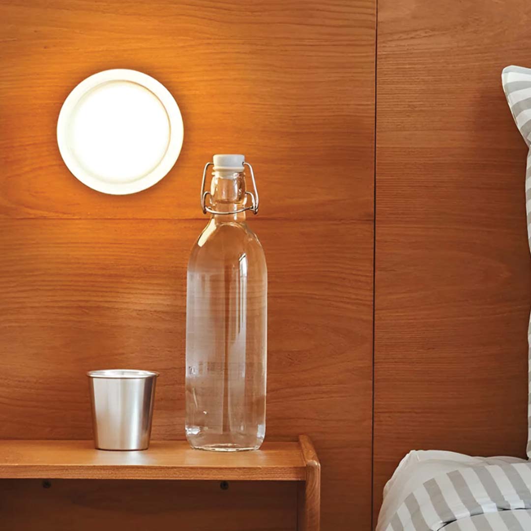 Een LichtAura wandlamp straalt een warme gloed uit boven een nachtkastje met een glazen fles en metalen beker, tegen een houten wand. Perfect voor een gezellige en moderne slaapkamerinrichting.