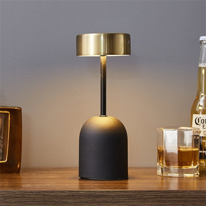 Lumeon LED-lamp met zwarte voet en gouden kap, geplaatst op een houten tafel. De lamp straalt warm, sfeervol licht uit en heeft een strak, modern ontwerp. Perfect als tafellamp voor woon- of slaapkamer. Energiezuinig en stijlvol.