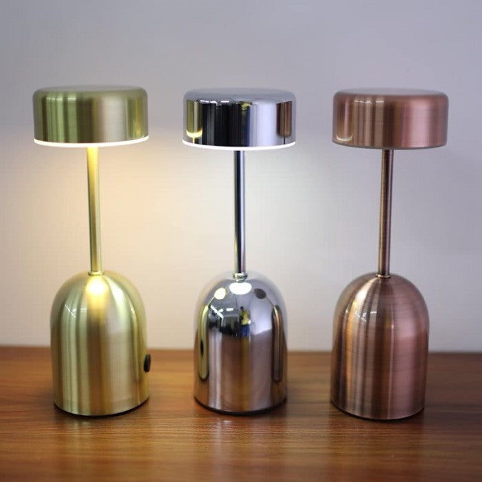 Drie Lumeon LED-lampen in goud, zilver en koper, staand op een houten oppervlak. De lampen hebben een strak en modern ontwerp met een cilindervormige kap en een smalle voet. Ideaal voor sfeerverlichting en energiezuinig in gebruik. Perfect als decoratief element in elk interieur.