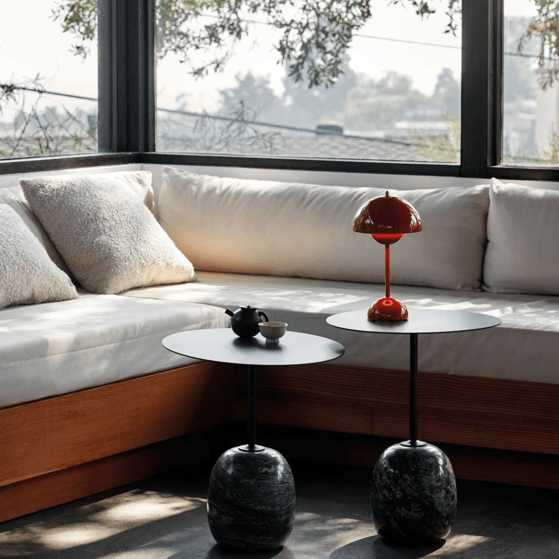 De LumiNova LED-tafellamp in een levendige rode kleur, elegant gepresenteerd op een modern bijzettafeltje naast een uitnodigende loungehoek met zachte kussens en een panoramisch uitzicht.