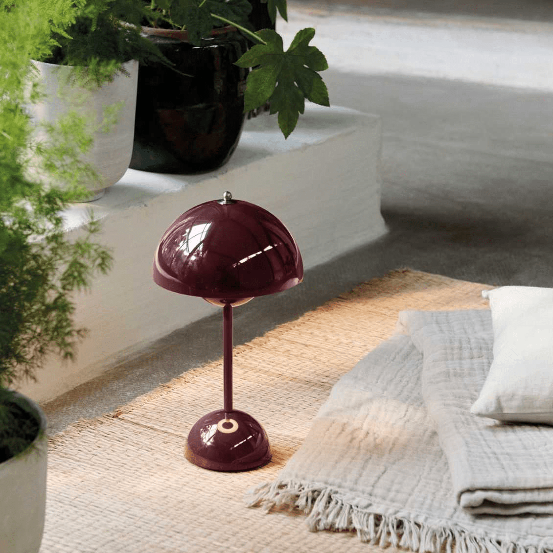 Dieprode LumiNova LED-tafellamp geplaatst op een neutraal vloerkleed, omringd door groene planten en zachte textielaccenten, creëert een warme en stijlvolle ambiance in uw interieur.