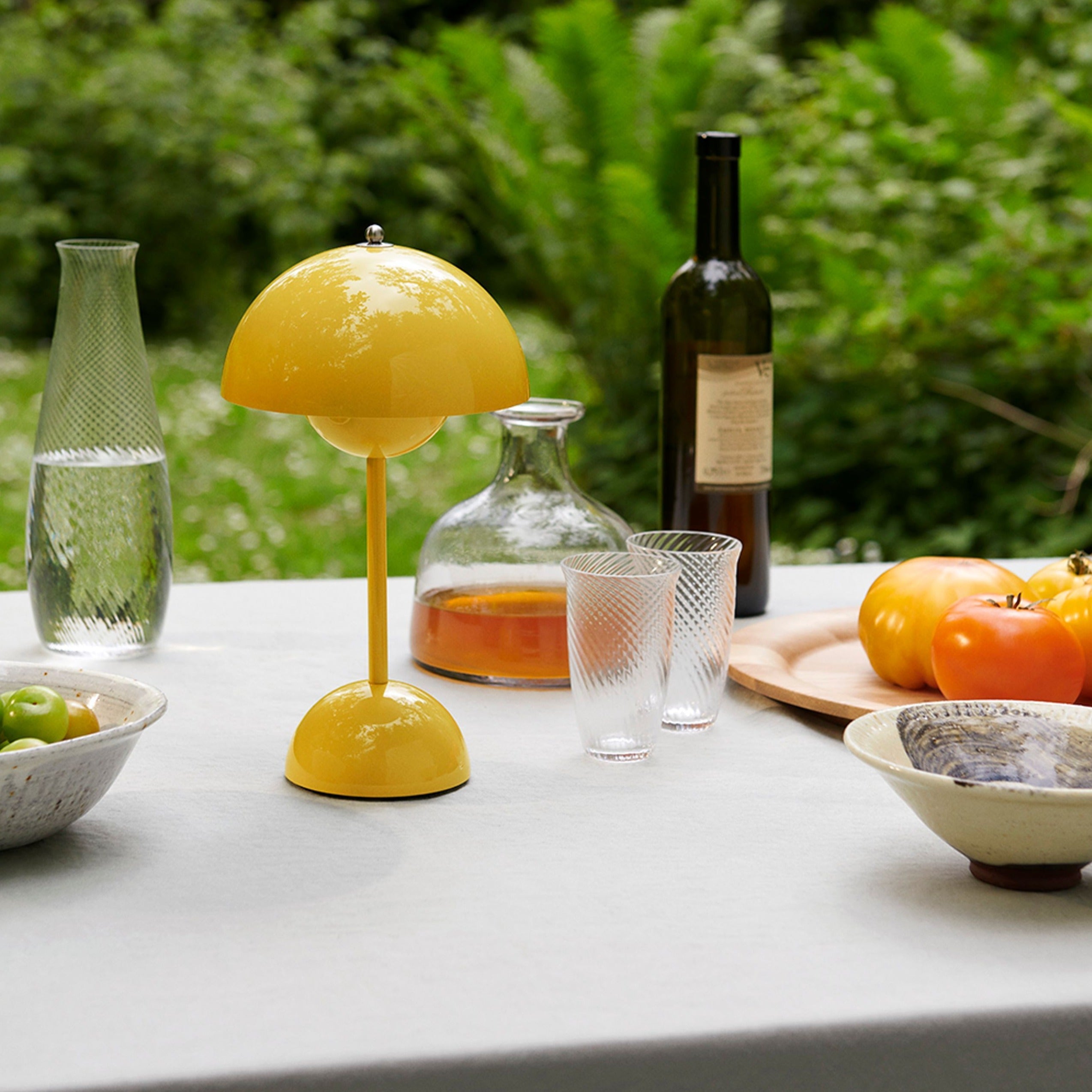 Een zonnig gele LumiNova tafellamp met LED-verlichting, geplaatst op een elegante buitentafel, omringd door glazen karaffen, borden met verse groenten en glazen. Deze stijlvolle lamp voegt een speels en modern accent toe aan elke buiten- of binnenruimte, ideaal voor sfeervolle verlichting.