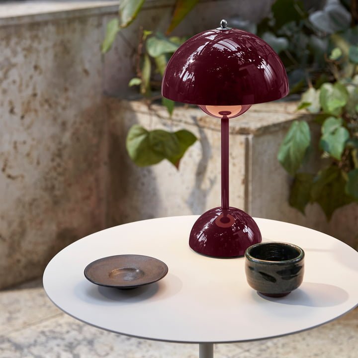 Een stijlvolle LumiNova tafellamp met een rijke bordeauxrode kap, geplaatst op een moderne tafel naast een zwart keramisch bord en een groene plant. De lamp verspreidt een zacht licht, ideaal voor het creëren van een gezellige en verfijnde sfeer in elke ruimte.