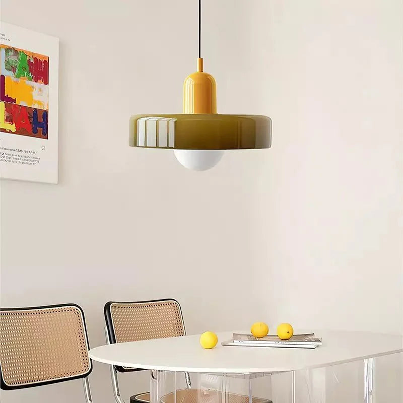 De Luminezza Living hanglamp in kleurrijk design hangt boven een minimalistische witte eettafel, omlijst door stijlvolle rotan stoelen. De geelgroene kap en speelse tinten van de lamp voegen een warme en moderne sfeer toe aan de ruimte, perfect voor een eigentijdse eetkamer.