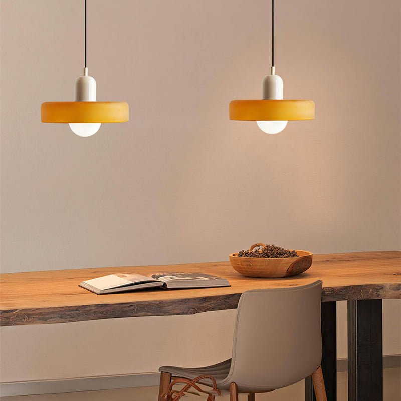 Twee Luminezza Living hanglampen in een kleurrijk design met warme gele kappen en elegante witte fittingen. Ideaal om een gezellige en stijlvolle ambiance te creëren boven een houten tafel in een moderne eet- of werkruimte.