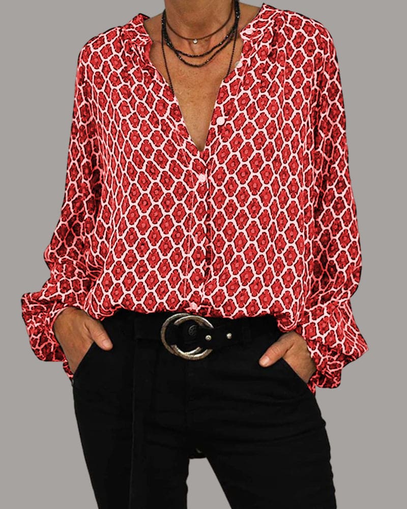 Een model draagt een elegante blouse met een rood-wit grafisch patroon, gecombineerd met een zwarte broek en een brede riem. De blouse heeft een V-hals en de look wordt afgemaakt met meerdere kettingen voor een verfijnde en trendy uitstraling.