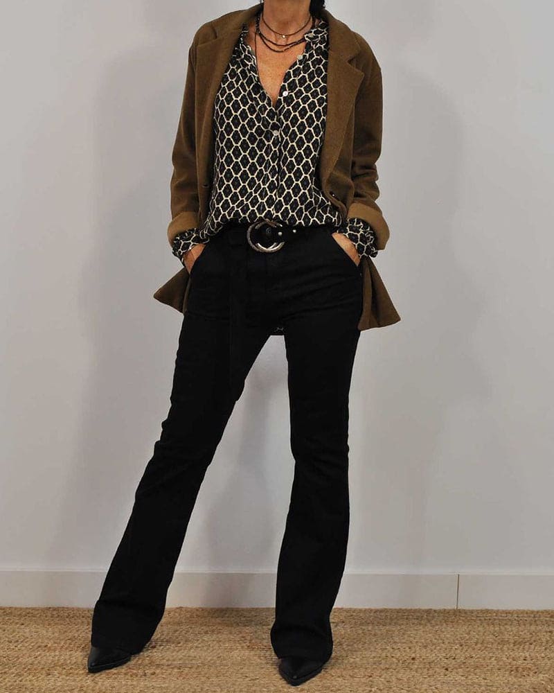 Een model draagt een stijlvolle blouse met een grafisch patroon in zwart en wit, gecombineerd met een bruine blazer en zwarte flare jeans. De outfit wordt afgemaakt met een brede riem en een chique ketting, wat een elegante en moderne uitstraling geeft.