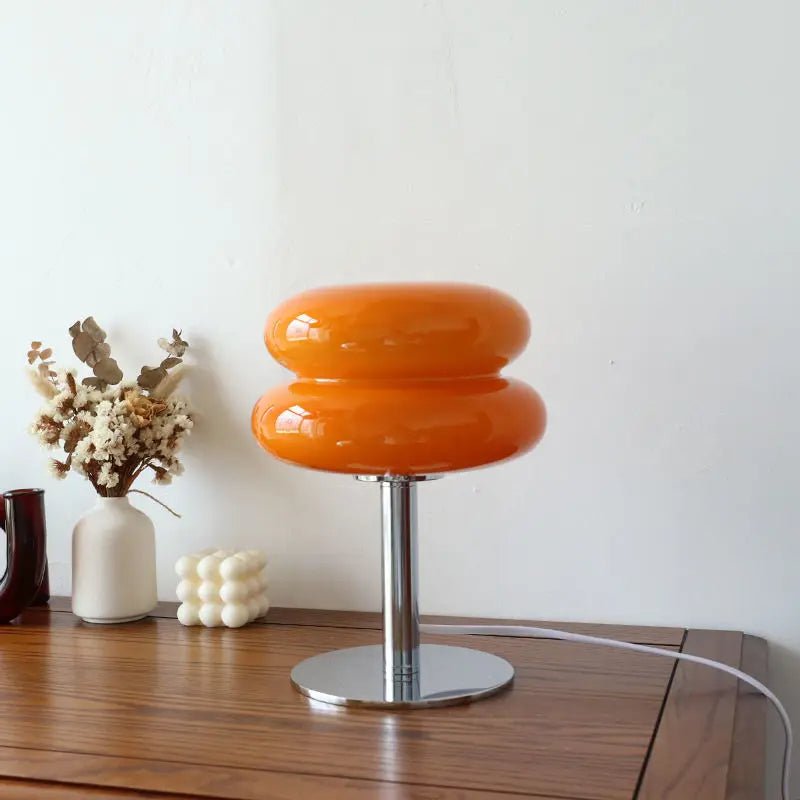 Iconische Luminore Design tafellamp in een opvallende oranje tint, met een dubbele glazen kap en een glanzende metalen voet. Geplaatst op een houten tafel met stijlvolle decoratieve accenten, perfect voor het creëren van een warme en moderne sfeer in elk interieur.