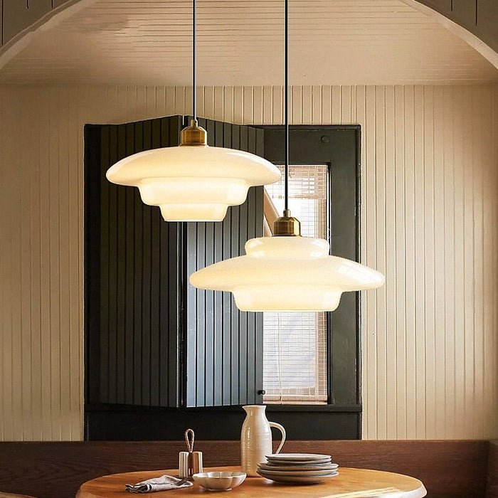 Alt text: A pair of elegant white glass pendant lamps with a unique layered design, suspended above a dining table. The lamps are complemented by warm lighting, creating a cozy and stylish atmosphere in the room. The light fixtures feature a brass accent at the top, adding a touch of sophistication to the modern interior design.