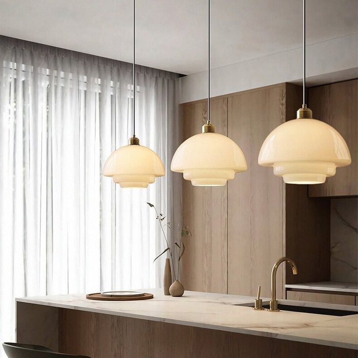 Three elegant white glass pendant lamps hanging over a modern kitchen counter, each featuring a smooth, layered design with a brass fixture at the top. The soft, warm glow from the lamps enhances the contemporary atmosphere, providing stylish and functional lighting. The pendant lights are suspended from black cords, creating a minimalist yet sophisticated look in the space.