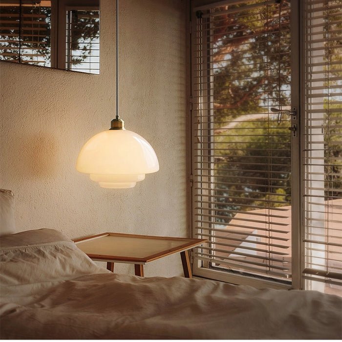 Alt text: A stylish white glass pendant lamp hanging in a bedroom, casting a warm, inviting glow. The lamp features a smooth, layered design with a brass fixture at the top, suspended from a black cord. The room is bathed in natural light from the window, with wooden blinds and a minimalist bed frame, creating a cozy and elegant ambiance.