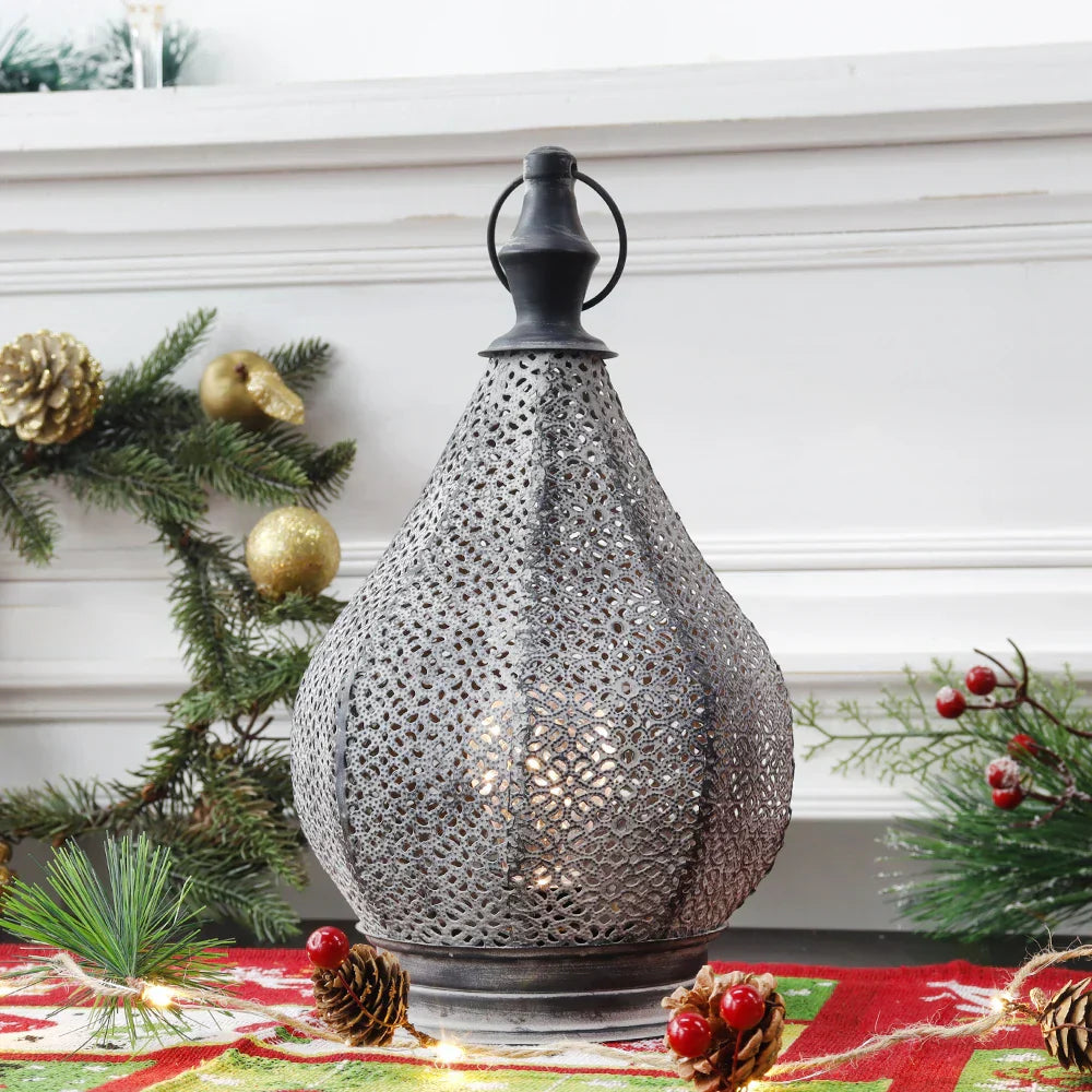 Een elegante tafel lamp met een fijn geperforeerd metalen ontwerp in de vorm van een waterdruppel. Het licht straalt zachtjes door de openingen in het metaal, waardoor een warme en gezellige gloed ontstaat. De lamp staat op een tafel, omgeven door kerstdecoraties, zoals dennenappels en takken, wat een feestelijke sfeer creëert. Perfect voor het toevoegen van een verfijnde en sfeervolle uitstraling aan elke ruimte.