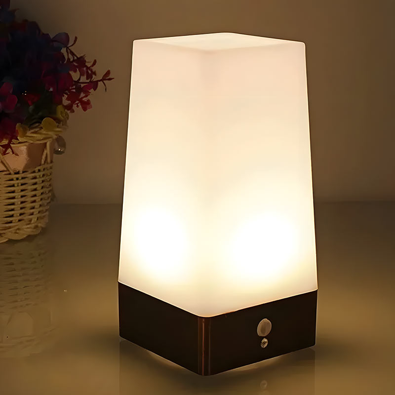 Luvora tafellamp met sfeervol diffuus licht, met een vierkante matglazen kap en een metalen basis. De lamp staat op een tafel in een gezellige setting, met een bloemenmand op de achtergrond. Ideaal voor nachtkastjes of woonkamers om een warme en uitnodigende sfeer te creëren.