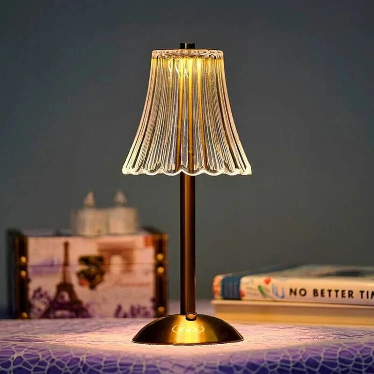 A modern, elegant wireless table lamp with a translucent pleated shade and a brass-colored base, glowing softly in a cozy setting with a book and decorative items in the background. Ideal for adding a touch of sophistication to any room.