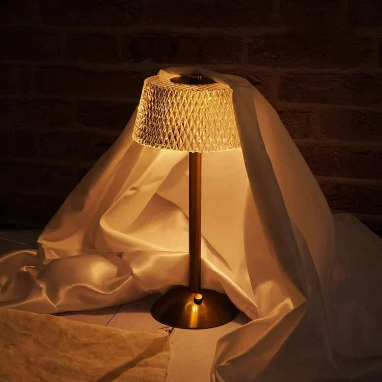 Een elegante draadloze tafellamp met een getextureerde lampenkap met een diamanten patroon, die een warme gloed uitstraalt over een met stof bedekte oppervlakte. De lamp heeft een elegante bronzen basis en voegt een moderne en verfijnde touch toe aan elke ruimte.